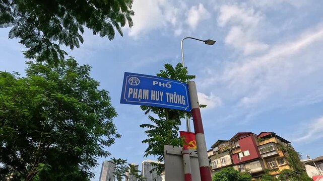 Blue street name sign of Pham Huy Thong street in Hanoi, Vietnam, with trees and city buildings under a blue sky.