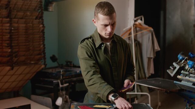 Medium shot of Caucasian male worker in coverall uniform sorting color palettes in screen printing and branding workshop, preparing materials for production process