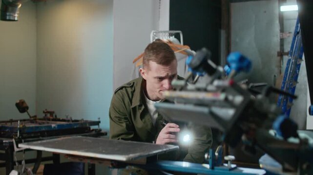 Medium shot of worker using flashlight to inspect machinery in screen printing and logo branding workshop