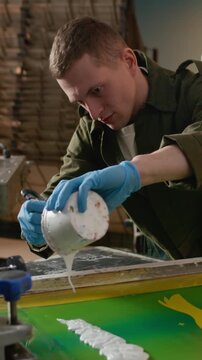 Vertical medium shot of male designer pouring white ink across bird design screen frame in workshop