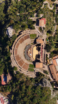 Aerial view of the Ancient Theatre of Taormina featuring the semicircular stone seating and historic ruins surrounded by lush green vegetation on a hillside in Taormina, Sicily, Italy.