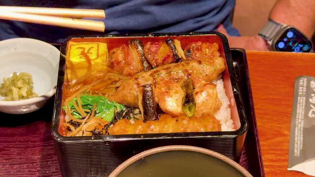 Person using chopsticks to eat japanese yakitori chicken bento box at restaurant.