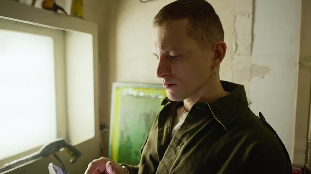 Dolly in medium closeup shot of young Caucasian man using smartphone while standing in branding workshop with screen printing equipment and lightbox, showing creative workflow and production process