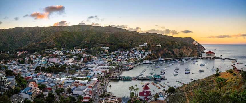Panoramic coastal town at sunset with hillside buildings, a curved harbor full of boats, and a prominent round waterfront landmark.