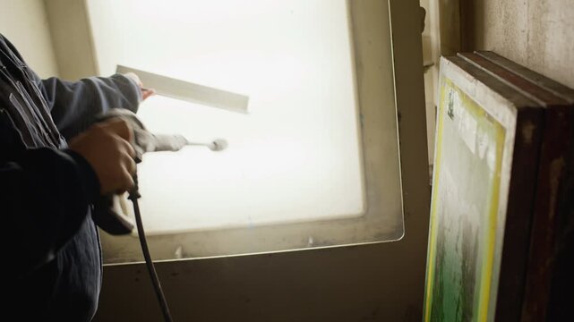 Close up shot of unrecognizable worker using spray gun to apply branding design or coating onto surface under bright backlight in workshop