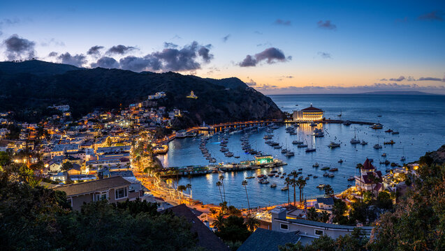 Coastal town at dusk with a curved harbor full of boats, warm building lights, surrounding hills, and a prominent round waterfront landmark.