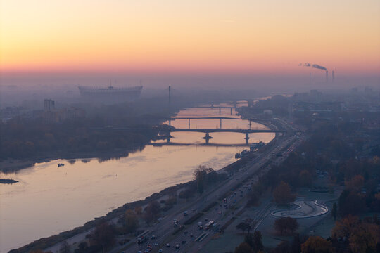 Aerial view of the Vistula River with bridges and the PGE Narodowy stadium under a hazy sunrise sky in Warsaw, Masovian Voivodeship, Poland.