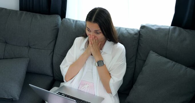 Surprised woman with laptop update. Woman sitting on couch experiences surprise from laptop notification. Seated woman displays joy and shock upon receiving unforeseen message on her computer