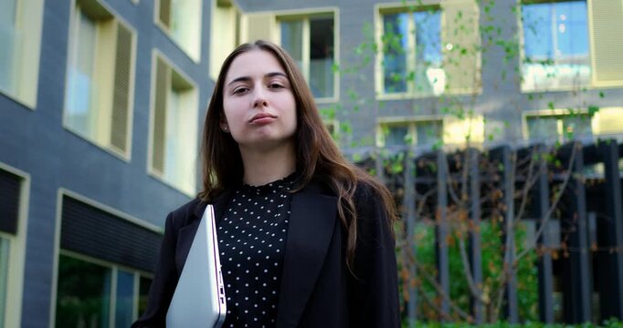Confident woman working. Determined businesswoman in polka dot blouse with laptop outside structure. Selfassured female entrepreneur holding her device near contemporary office environment