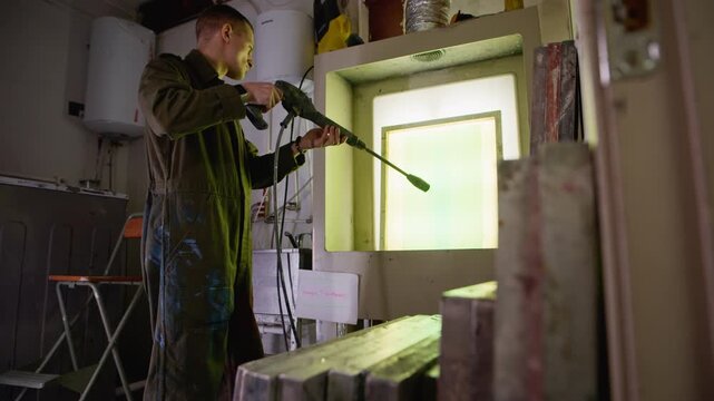 Medium wide shot from behind of worker wearing coveralls applying coating with spray gun to printing frame in workshop