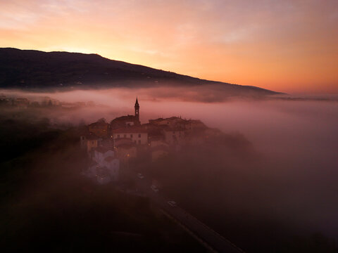 Aerial view of the historic village of Piantravigne emerging from a thick layer of morning mist with its church tower silhouetted against a vibrant sunrise sky Piantravigne, Tuscany, Italy.