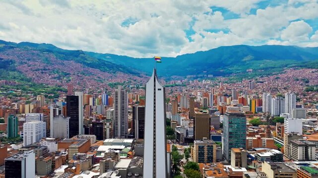 Aerial drone view of downtown Medell&iacute;n, Colombia, featuring a waving flag atop a skyscraper above the dense urban skyline and surrounding cityscape in clear daylight.