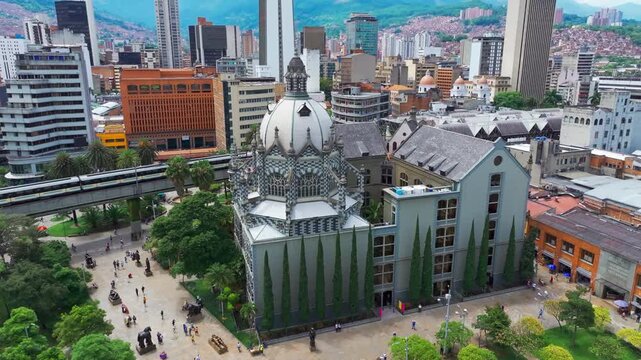 Aerial drone view of Botero Square in Medell&iacute;n, Colombia, with the metro passing nearby, showcasing the public plaza, urban architecture, and dynamic city atmosphere from above.
