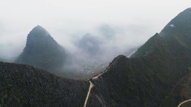 Aerial footage of a mist covered mountain valley with a hidden rural village barely visible beneath low clouds, surrounded by rugged karst peaks creating a moody cinematic highland landscape