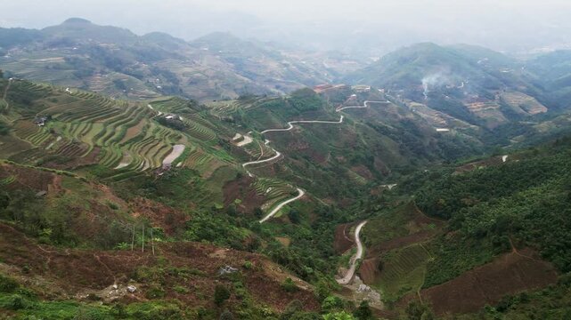 Aerial drone view of highland landscape with layered rice terraces and winding roads cutting through cultivated farmland across rolling hills under soft haze and moody weather in Ha Giang Vietnam