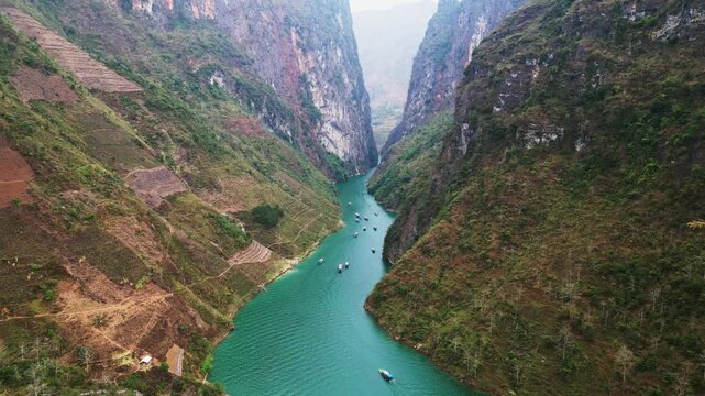 Cinematic aerial view of winding river flowing through a limestone canyon with towering rock walls terraced farmland and tourist boats exploring a deep mountain gorge in Ha Giang Vietnam