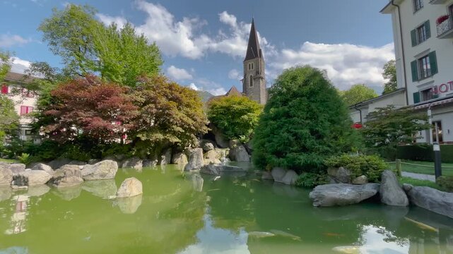 Static shot, partly cloudy sunny day, Japanese garden koi pond with church steeple backdrop in Interlaken, Switzerland. Serene and picturesque.
