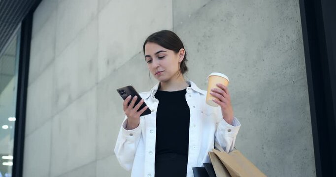 Young woman examining options outdoors. Casual shopper assessing selections amidst city storefronts. Modern woman thoughtfully comparing shopping choices while enjoying coffee outdoors