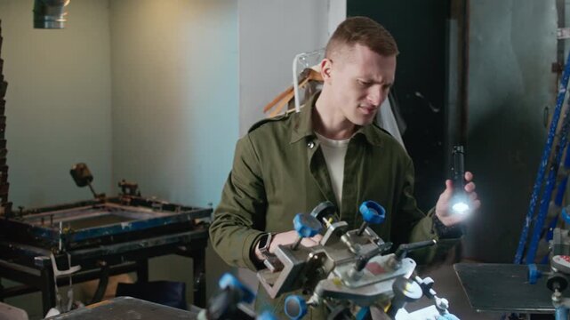 Medium shot of Caucasian male worker observing machines in printing workshop with flashlight used for screen printing and logo production