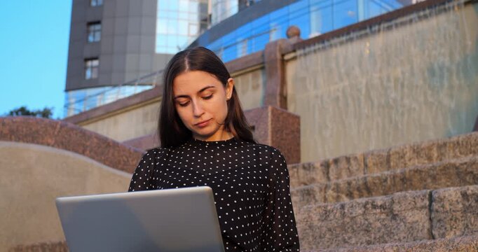 Casual woman on outdoor steps. Casual female relaxing on stone stairs with laptop and coffee. Woman leisurely sipping coffee while browsing laptop during relaxed outdoor evening break