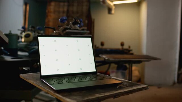 White screen laptop in sharp focus in foreground with rotating screen printing machine and frames in background of empty workshop