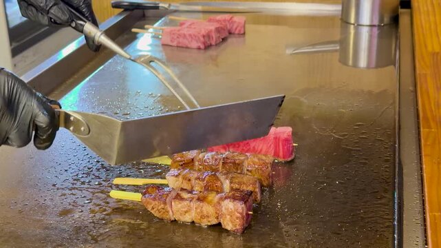 Professional chef grilling premium wagyu beef skewers on hot teppanyaki iron griddle with spatula.