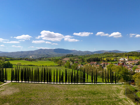 Aerial view of the Balze landscape with a long row of cypress trees, green fields, and a small village under a blue sky in Reggello, Tuscany, Italy.