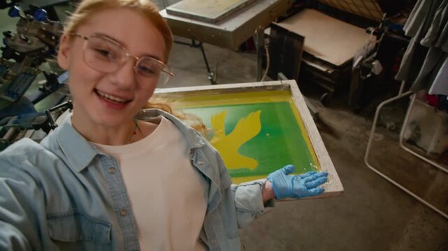 Handheld POV shot of female designer speaking to camera inside screen printing workshop with bird artwork on screen installed on production machine in background
