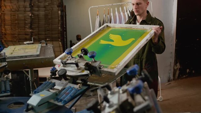 Medium shot of Caucasian male worker positioning screen printing frame with bird image onto machinery equipment for logo branding or screen printing production in workshop
