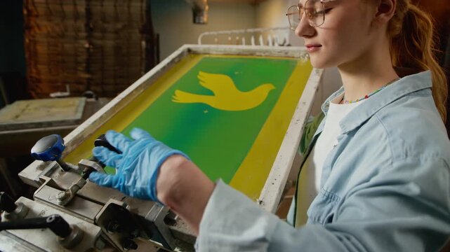 Stab medium side view shot of young female worker placing screen frame with bird artwork on machinery for logo branding and screen printing in workshop