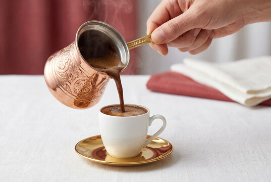 Hand pouring hot traditional turkish coffee from copper cezve into white cup on table
