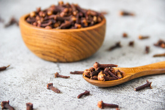 Dark brown cloves fill a wooden bowl. Scattered cloves rest on a gray concrete table. Wooden spoon holds a few cloves