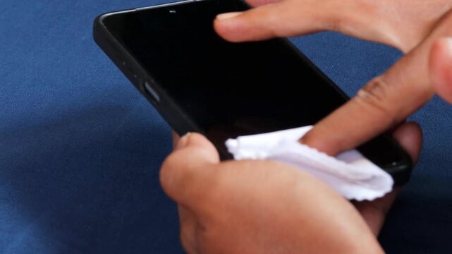 Close-up cropped shot of Asian man hand diligently disinfecting smartphone screen with microfiber cloth, preventing virus transmission and maintaining personal device hygiene.