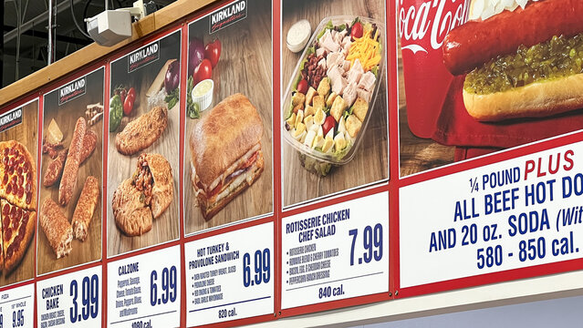 Menu board displaying various food items at Costco, featuring prices and calorie counts, with Kirkland brand prominently featured alongside Coca-Cola products