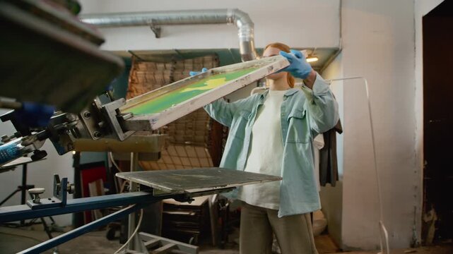 Side view stab medium shot of young Caucasian female worker mounting screen printing frame onto equipment for branding production process