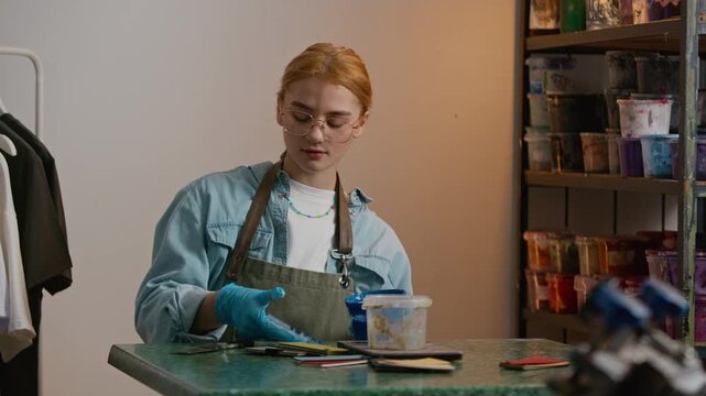 Stab waist up shot of young Caucasian female designer wearing gloves and apron reviewing paint jars and selecting colors for branding work in professional workshop