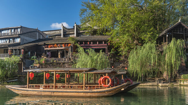 Beautiful wooden tourist boat by the river bank. Red traditional lanterns are suspended. Lifebuoys on board. Green trees, buildings against a background of blue sky and clouds. China. Fenghuang 
 