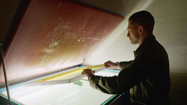 Side view eye level medium shot of young Caucasian male artist interacting with illuminated screen printing table during branding work in dimmed lit workshop