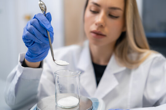 Conducting accurate measurement of powdered substances with a stainless steel spoon at laboratory workstation. Clean and sterile environment with visible analytical tools and beaker present.