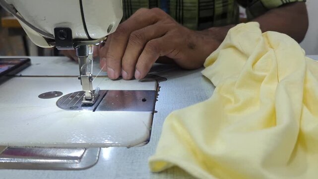 Close-up of a professional tailor's hands meticulously threading a needle on an industrial sewing machine before starting to stitch a yellow garment in a textile workshop.