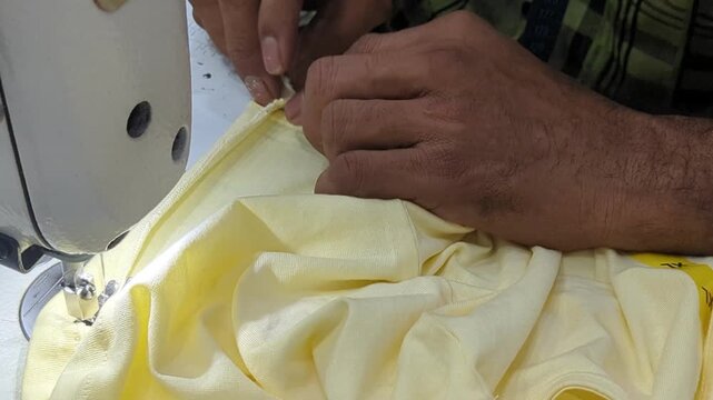 Close-up of professional seamstress hands operating an industrial sewing machine to stitch the hem of a yellow cotton garment in a textile factory.