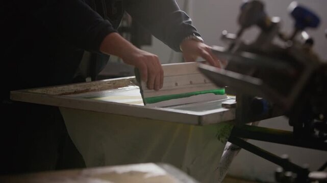 Cropped shot of unrecognizable male worker using squeegee to push ink over screen frame on printing machine in workshop, showing precision and branding production process