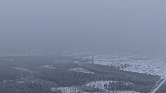 Drone ascending into fog above a large wind energy landscape in winter conditions. Multiple turbines spread across snowy terrain gradually fade into low clouds and distant horizon.