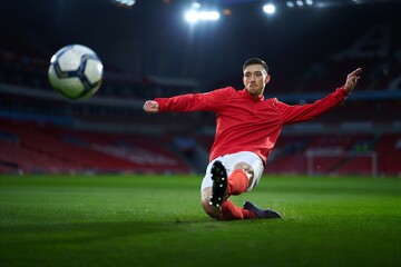Naklejka premium Athletic young man in red jersey performing a dynamic soccer kick on a green grass field with stadium lights overhead