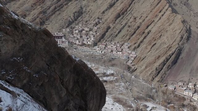 Hemis Monastery sits nestled in a steep Himalayan valley surrounded by barren mountain slopes and traditional buildings. Forward tracking aerial shot descends toward the Buddhist monastery complex.