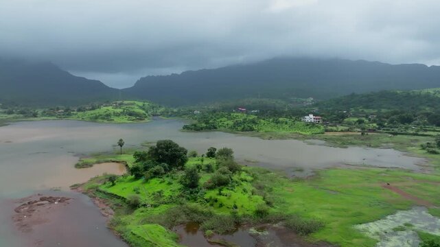 Post-monsoon drone footage panning across verdant Maharashtra landscape featuring water body with islet, agricultural lands, hill ranges, and rural settlement near Vikatgad Peb Fort.