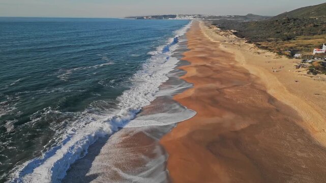 Cinematic aerial drone shot of the long sandy coastline at Praia do Salgado in Nazare, Portugal. Blue Atlantic waves crash onto the shore under a clear sky