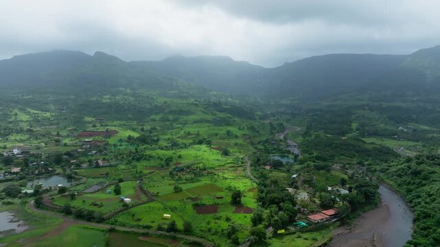 Maldunge, Maharashtra's lush green valley unfolds below Vikatgad Peb Fort after monsoon rains. Slow forward aerial tracking shot reveals agricultural patchwork, scattered villages, and misty mountain.