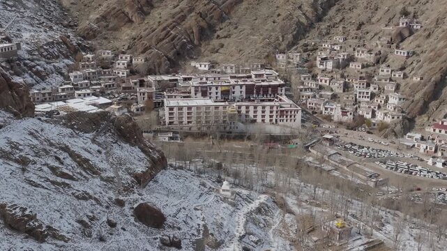 Hemis Monastery's white multi-story structure sits among traditional village buildings in a high-altitude Himalayan valley with snow-dusted slopes. Slow aerial shows Gompa complex with houses..