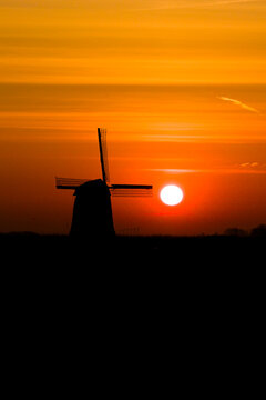 Aerial view of the silhouette of a traditional windmill against a glowing orange sunset sky with horizontal clouds in Schermerhorn, North Holland, Netherlands.